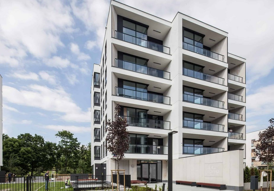 Nowoczesny budynek mieszkalny z balkonami i dużymi oknami, widok z zewnątrz, na pierwszym planie drzewo z brązowymi liśćmi.