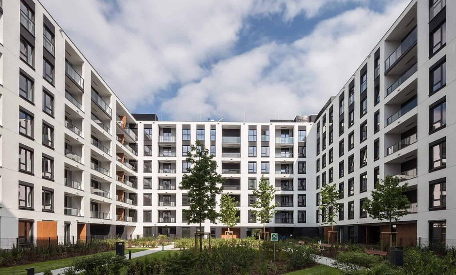 Nowoczesny dziedziniec osiedla mieszkaniowego z białymi budynkami i czarnymi ramami okiennymi, zielenią trawników i drzew, błękitnym niebem w tle.