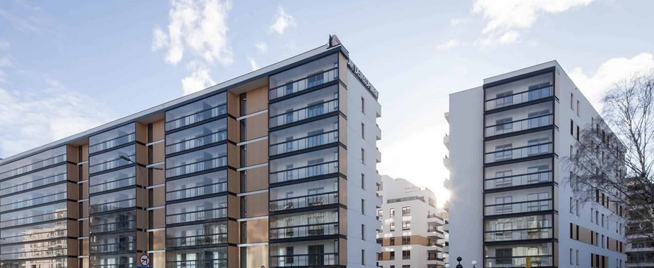 Nowoczesny budynek apartamentowy z przeszklonymi balkonami, widziany pod kątem, z logo dewelopera na dachu.