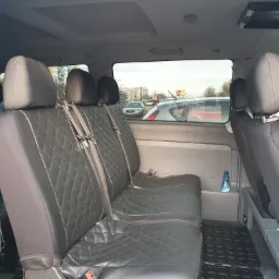 Wnętrze busa z czarnymi, pikowanymi siedzeniami, widok z tyłu na okno i zaparkowane samochody, niebieska butelka wody przy tylnym siedzeniu.