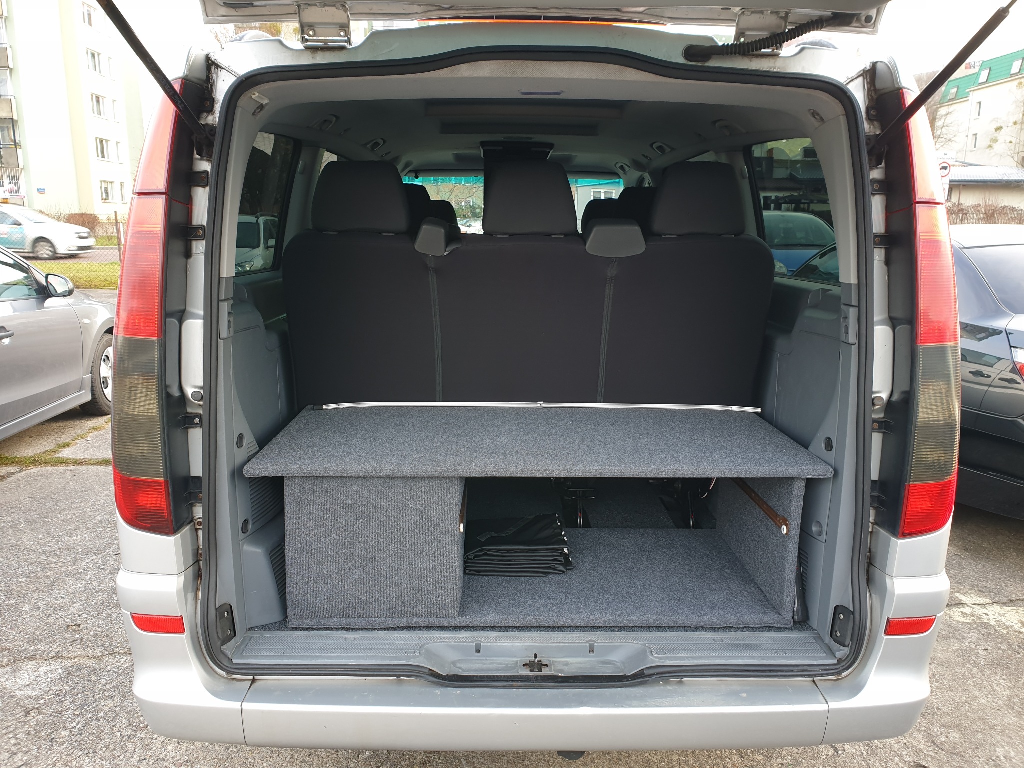 Widok z tyłu srebrnego vana z otwartą klapą bagażnika, ukazujący szarą zabudowę bagażnika z półką i schowkiem, prawdopodobnie dla celów turystycznych lub kempingowych.