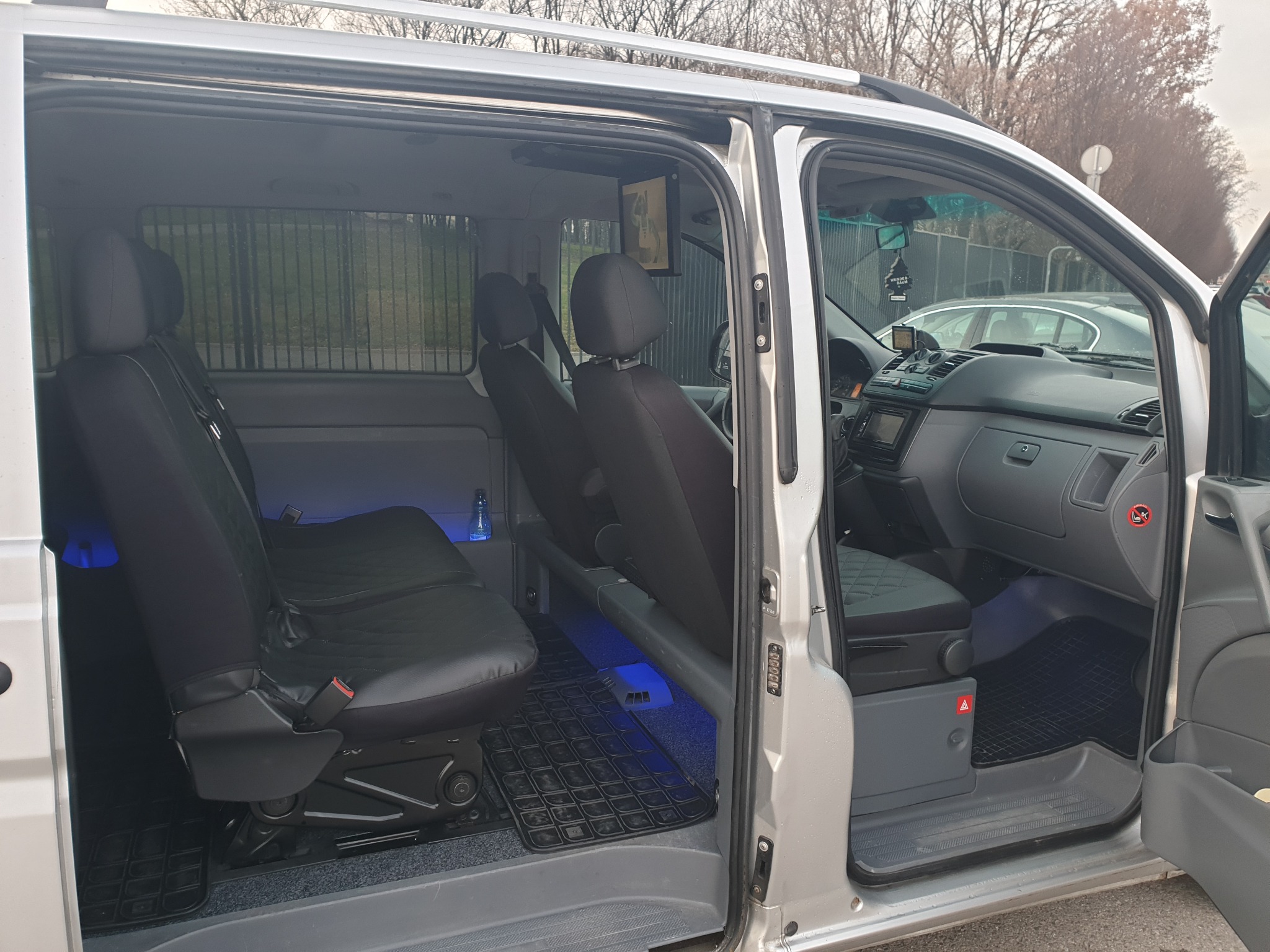 Wnętrze srebrnego busa z trzema rzędami siedzeń, widoczne czarne pokrowce na siedzeniach, podświetlenie LED pod siedzeniami i monitor na ścianie.