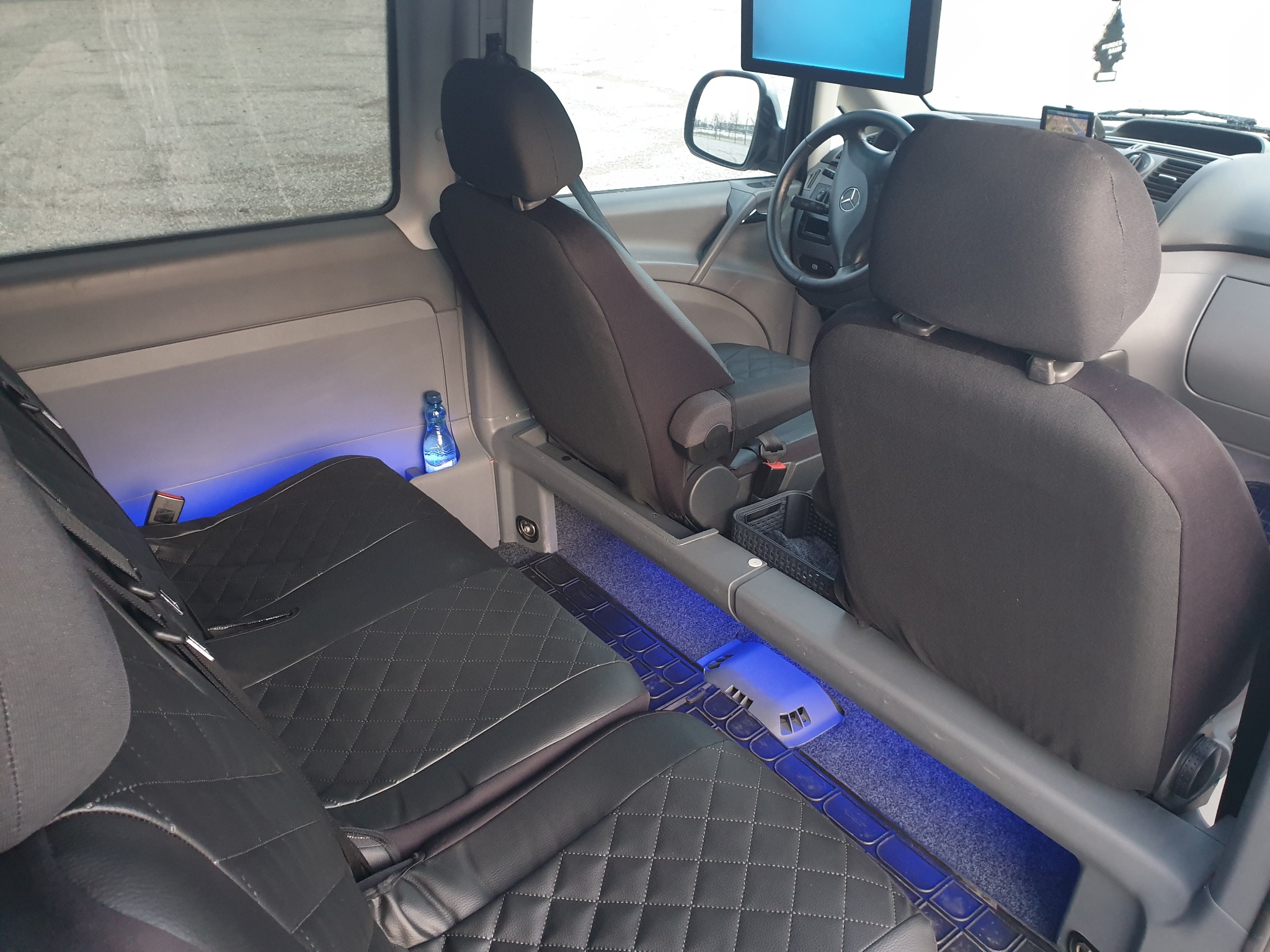 Wnętrze busa z czarną tapicerką w romby, podświetleniem LED w kolorze niebieskim, oraz zamontowanym monitorem dla pasażerów.