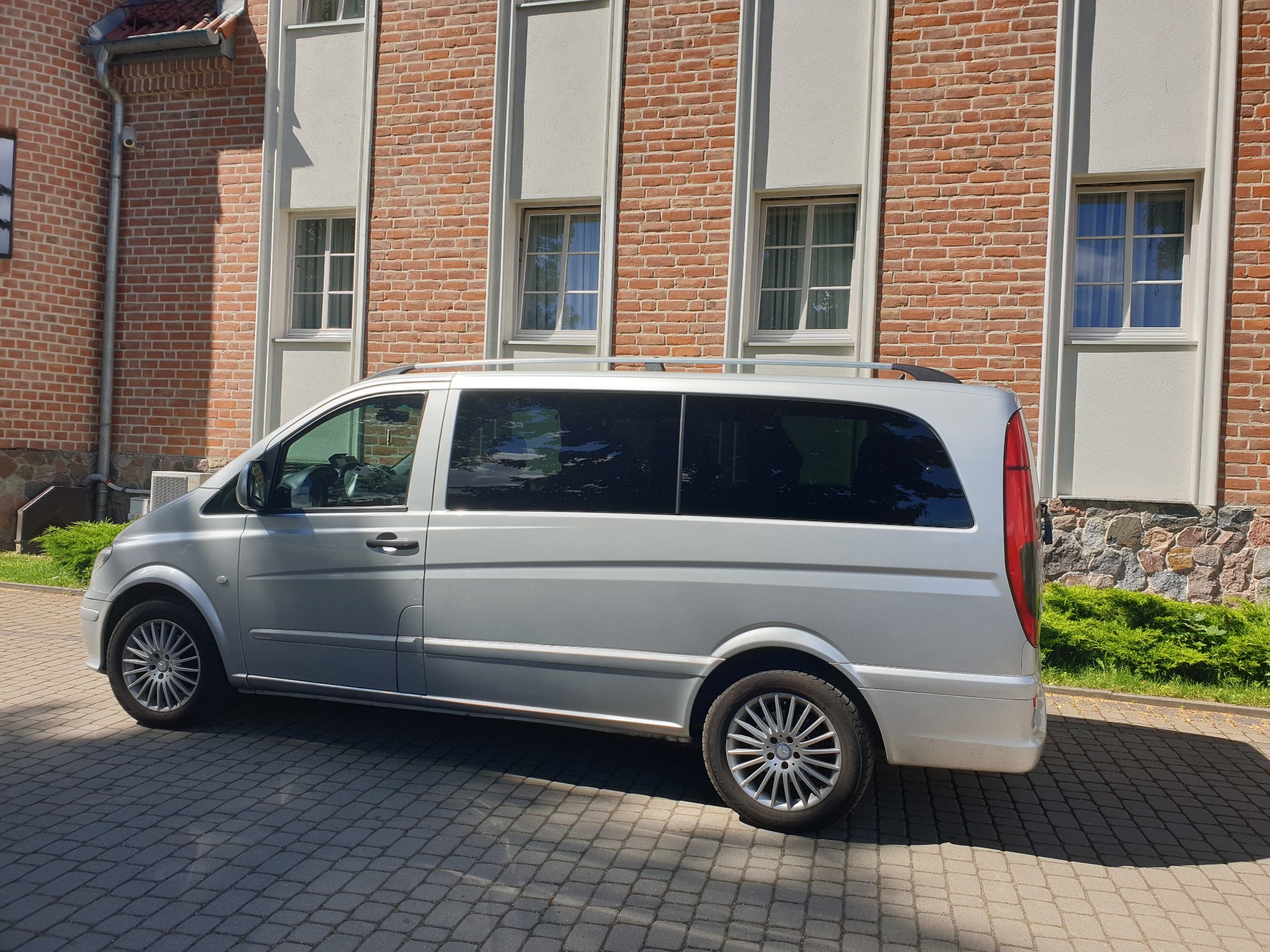 Srebrny van Mercedes-Benz Vito zaparkowany przed budynkiem z czerwonej cegły z jasnymi obramowaniami okien.