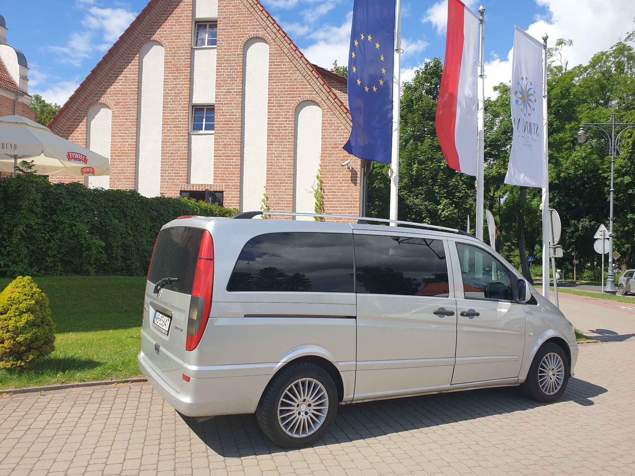 Srebrny van Mercedes Viano zaparkowany na brukowanym placu, z flagami UE i Polski oraz budynkiem w tle.