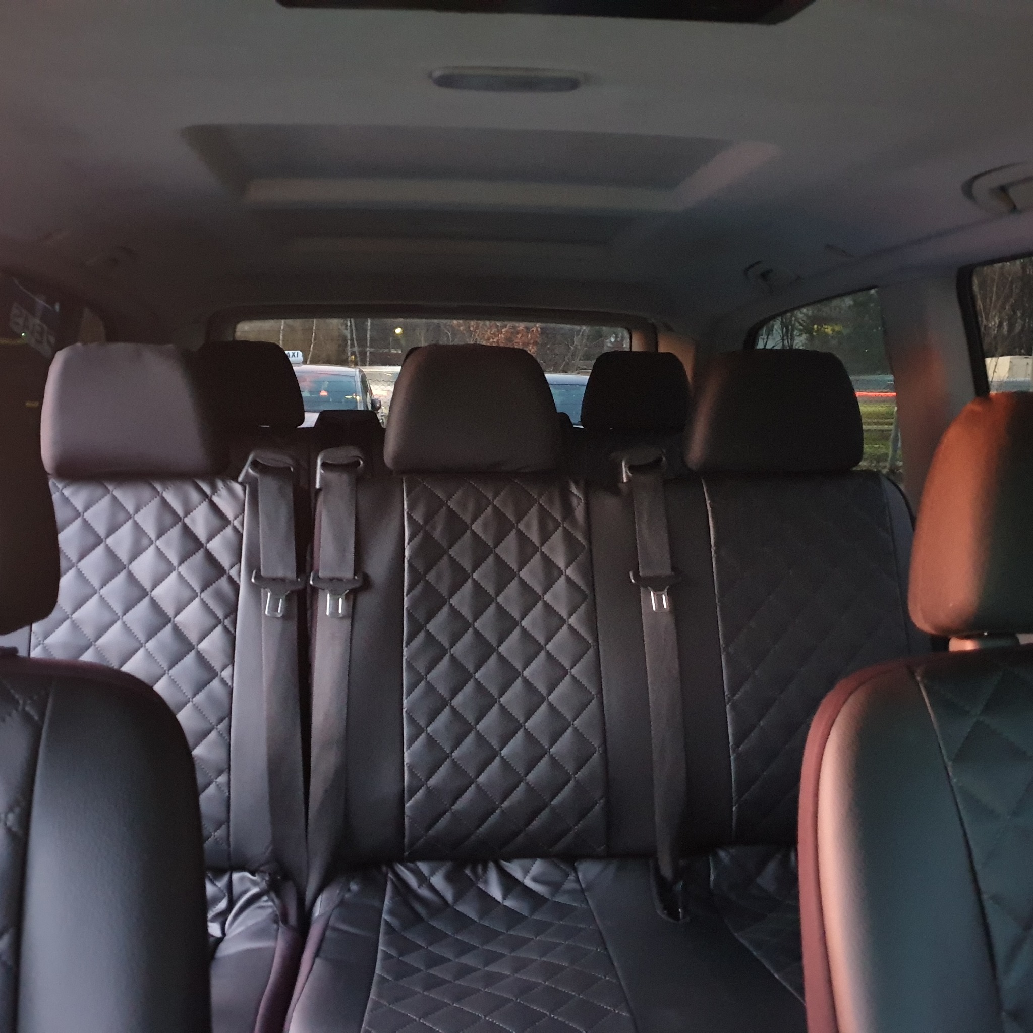 Wnętrze samochodu z czarną, pikowaną tapicerką siedzeń pasażerskich z tyłu, widoczne pasy bezpieczeństwa i zagłówki, w tle widok na ulicę i taksówkę.