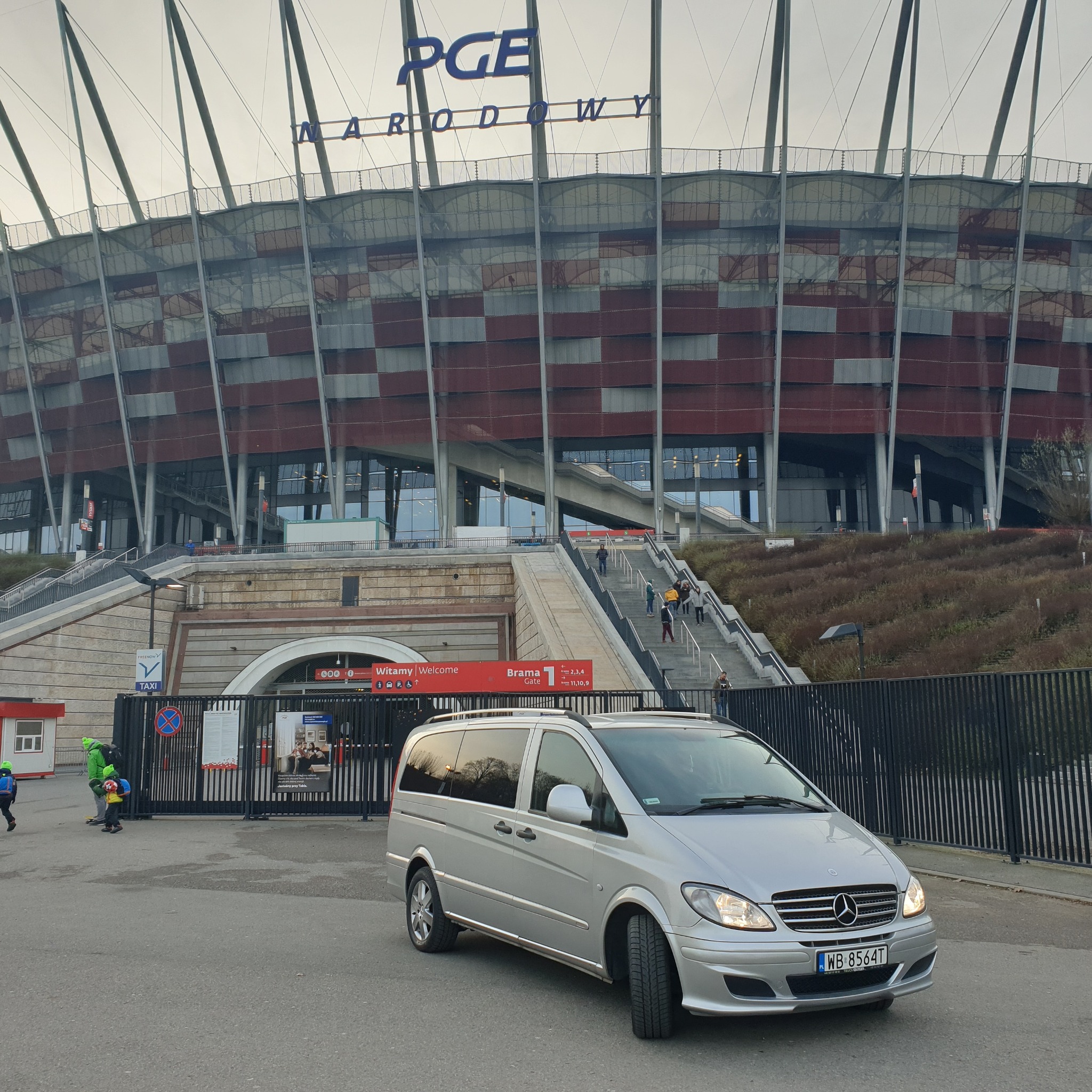 Srebrny Mercedes Viano zaparkowany przed PGE Narodowym w Warszawie, widoczny fragment bramy wejściowej i schody z przechodniami.