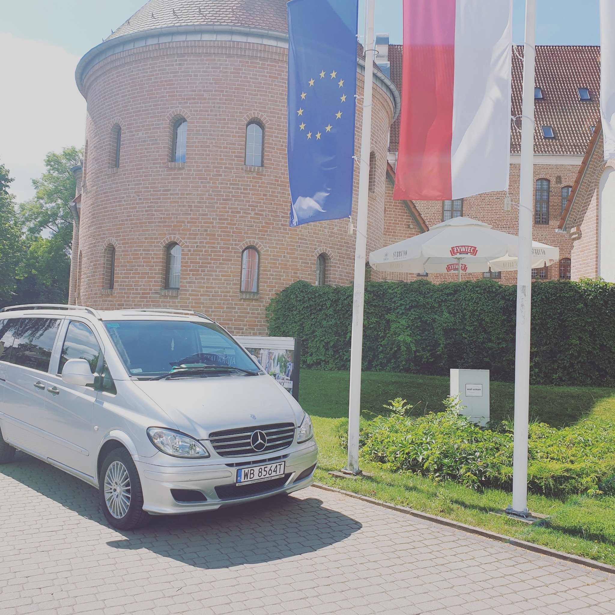 Srebrny Mercedes Vito zaparkowany na brukowanym placu przed ceglanym budynkiem z wieżą, z flagami Polski i Unii Europejskiej w tle.