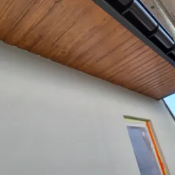 Podbitka dachowa z drewnopodobnym wzorem, zamontowana przy szarej elewacji budynku w Nowym Targu, widoczna z dołu pod kątem, z fragmentem orynnowania i rusztowania.