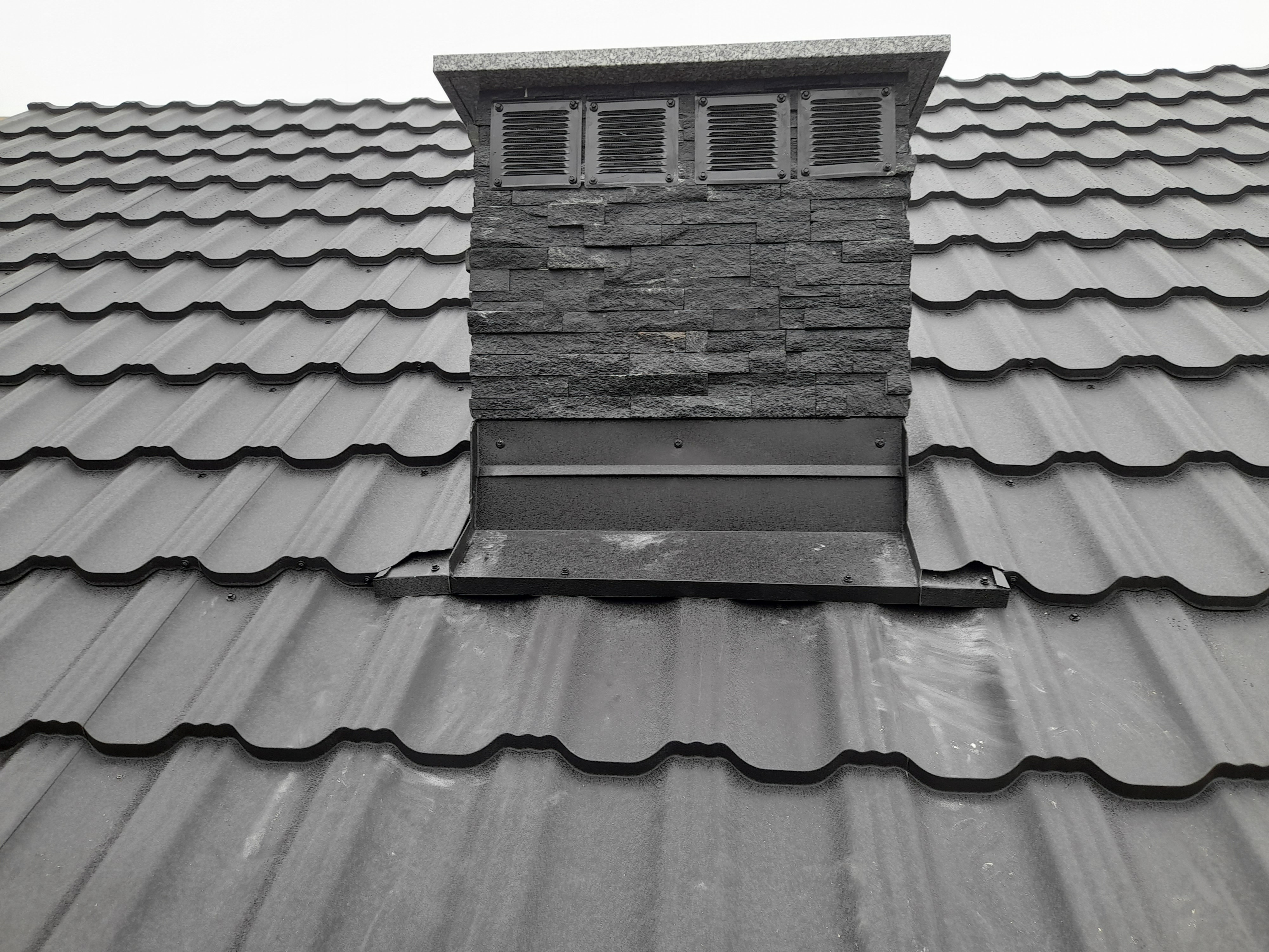 Szary dach z blachy trapezowej z kominem wykończonym kamieniem i kratkami wentylacyjnymi.