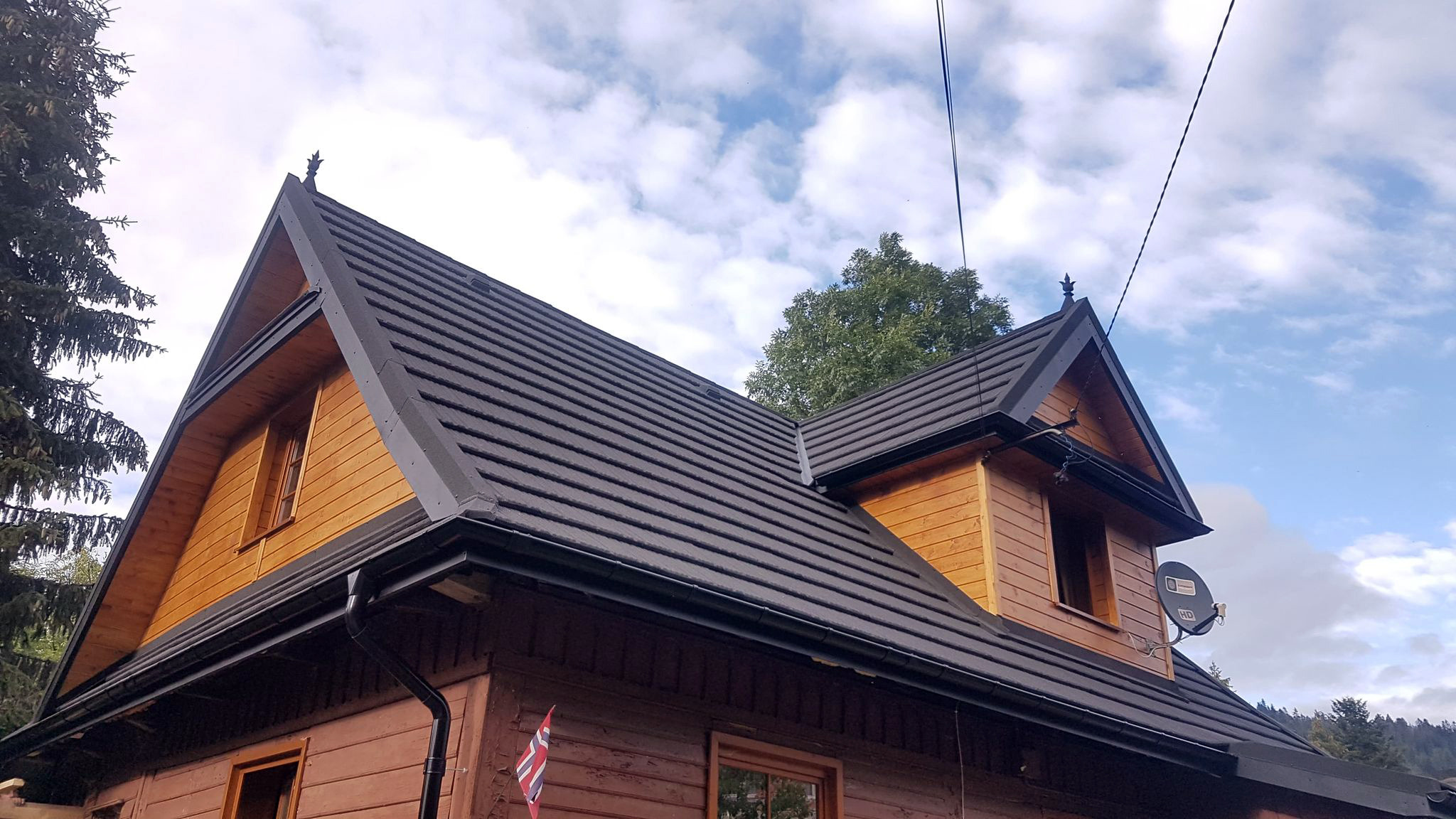 Nowy dach z blachodachówki modułowej na drewnianym domu w Nowym Targu, widok z dołu na tle błękitnego nieba z chmurami.