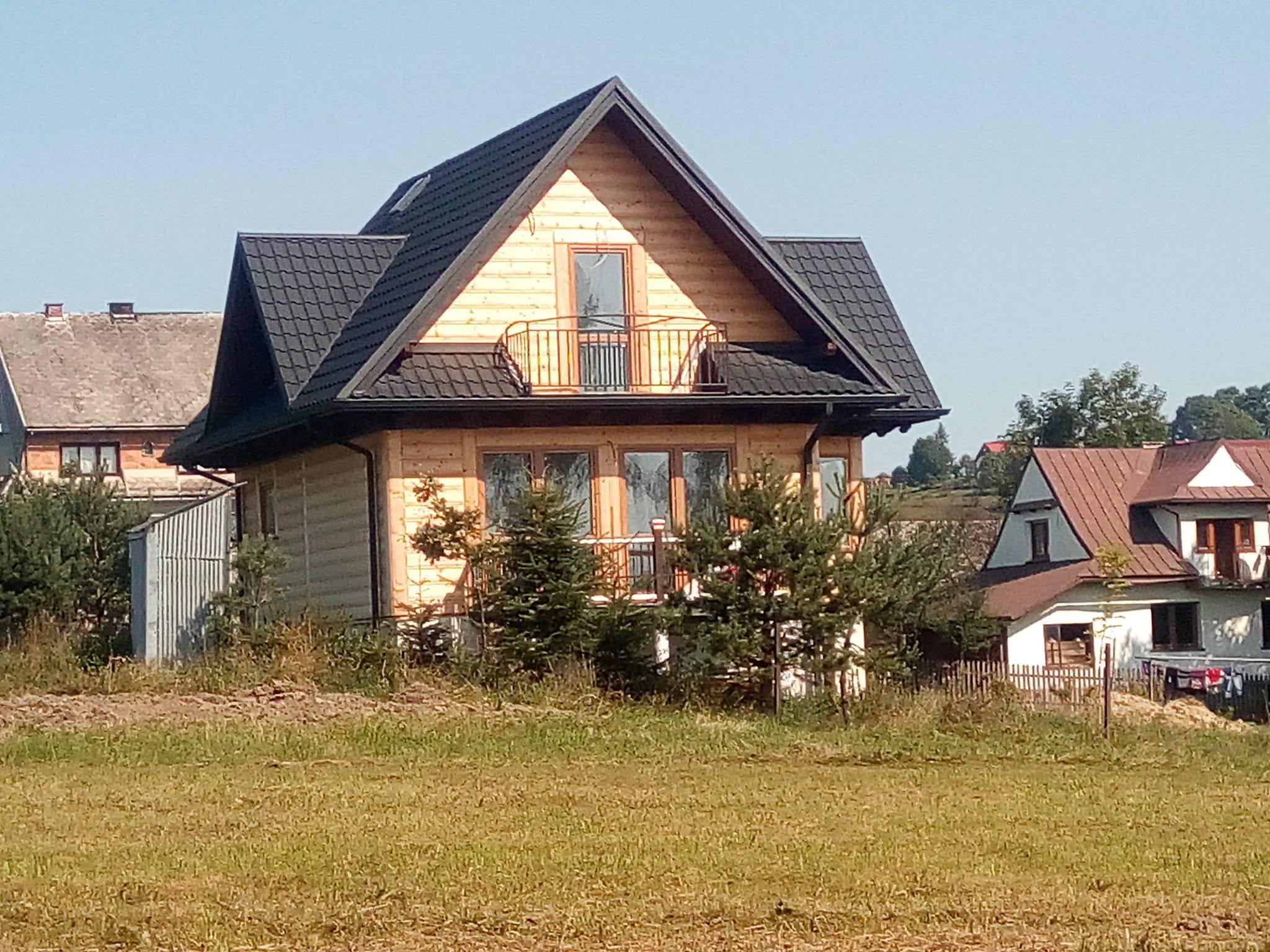 Nowoczesny dom drewniany z ciemnym, metalowym dachem dwuspadowym i małym balkonem na poddaszu, otoczony młodymi drzewkami iglastymi na tle łąki i innych budynków.