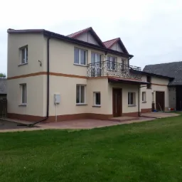 Dwukondygnacyjny dom z beżową elewacją, brązowymi akcentami i balkonem z kutą balustradą, widoczny z perspektywy trawnika w pochmurny dzień.