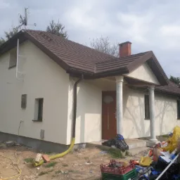 Dom jednorodzinny po świeżym tynkowaniu elewacji w kolorze kremowym, widoczne ślady prac wykończeniowych na posesji, brązowa dachówka i ceglany komin, antena satelitarna na dachu.