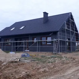 Dom jednorodzinny w trakcie prac elewacyjnych, widoczne rusztowania, ocieplenie styropianem i nowa dachówka w kolorze grafitowym, przygotowany teren wokół budynku.
