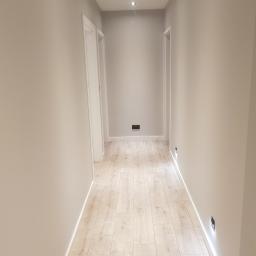 Bilders. - Wąski korytarz w odcieniach szarości z panelami podłogowymi imitującymi jasne drewno, oświetlony punktowo w suficie i w dolnej części ścian.