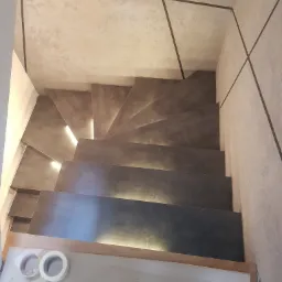 Spiralne schody z podświetleniem LED, wykończone w szarym betonie architektonicznym, widok z góry, taśma malarska na podłodze.