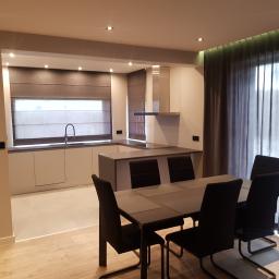 Bilders. - Nowoczesna kuchnia z jadalnią: minimalistyczne białe szafki, szare rolety rzymskie, stół z krzesłami na drewnianej podłodze, oświetlenie punktowe i zasłony.