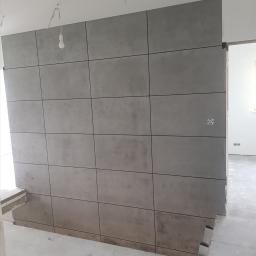 Bilders. - Ściana wykończona szarymi płytami wielkoformatowymi z fugami, widoczne niedokończone prace w pomieszczeniu, surowy beton na podłodze i wisząca żarówka bez klosza.