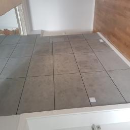 Bilders. - Ułożona, szara, matowa podłoga z płytek w pomieszczeniu w trakcie remontu, widoczne wystające kable elektryczne i fragment ściany w stanie surowym.