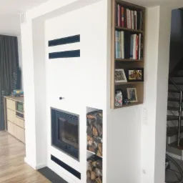 Nowoczesny kominek z wkładem, p&oacute;łką na drewno i wbudowaną biblioteczką w minimalistycznym wnętrzu. Widoczne czarne akcenty w postaci szczelin wentylacyjnych i obramowania kominka.