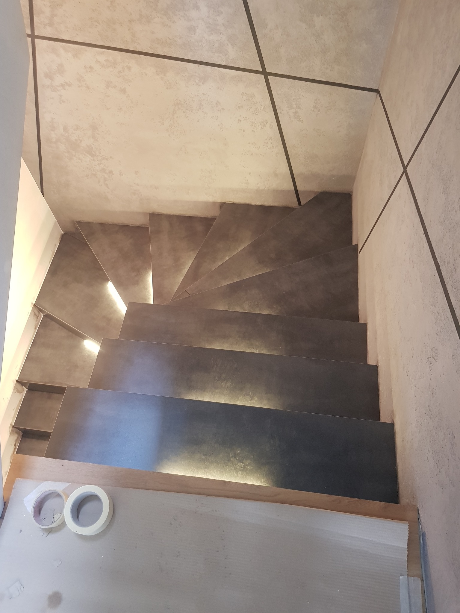 Spiralne schody z podświetleniem LED, wykończone w szarym betonie architektonicznym, widok z góry, taśma malarska na podłodze.