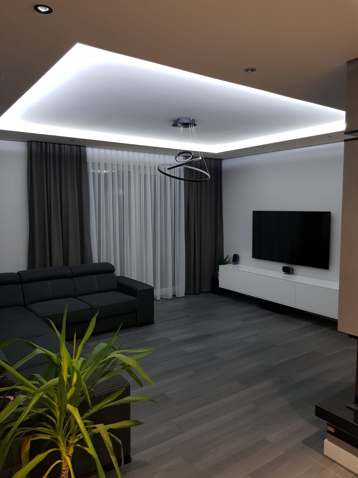 Nowoczesny salon z szarą sofą narożną, drewnianą podłogą w odcieniach szarości, białymi meblami RTV, designerską lampą sufitową i podświetlanym sufitem podwieszanym.