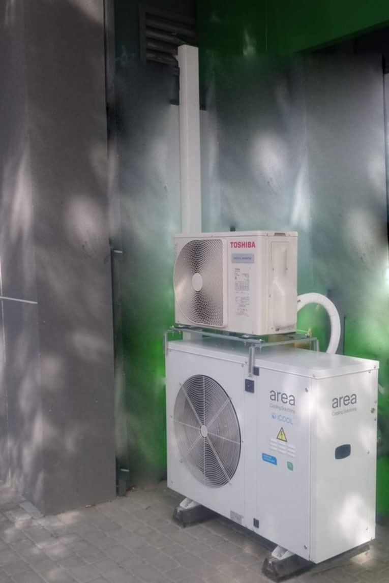 Dwie jednostki zewnętrzne klimatyzacji zamontowane jedna na drugiej na zewnątrz budynku, z widocznymi przewodami i elementami montażowymi, na tle ściany z efektem graffiti.