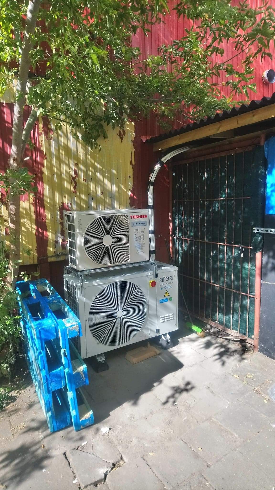 Dwa agregaty klimatyzacyjne Toshiba i Area Cooling Solutions ustawione jeden na drugim na paletach i drewnianych podkładkach przed pomalowaną na czerwono i żółto ścianą budynku z widoczną kratą...