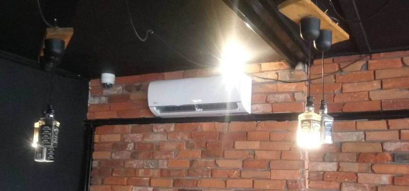 Biały klimatyzator ścienny zamontowany na ścianie z czerwonej cegły w pomieszczeniu z czarnym sufitem, obok lampa wisząca wykonana z butelki po whisky i kamera monitoringu.