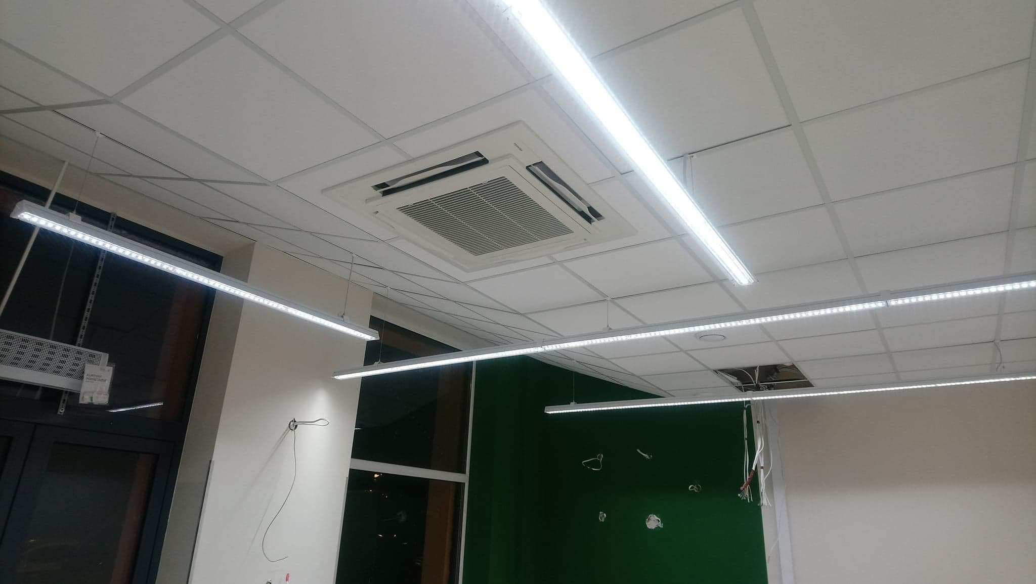 Sufit podwieszany z zamontowaną kasetą klimatyzacyjną i liniowymi lampami LED, widoczne przewody elektryczne w trakcie instalacji.