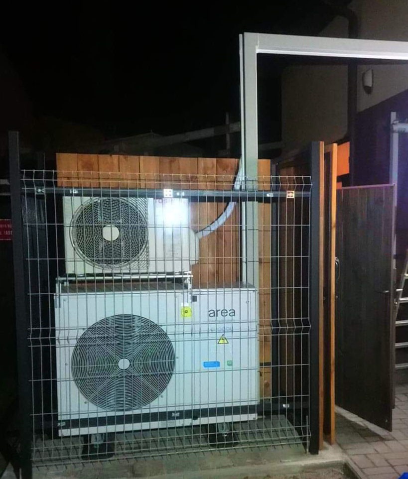 Dwie jednostki zewnętrzne urządzenia chłodniczego marki Area, umieszczone jedna nad drugą za metalową siatką ogrodzeniową, widoczne w słabym oświetleniu nocnym.