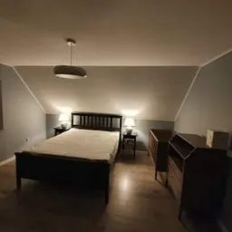 Pokój na poddaszu z drewnianą podłogą, czarnym łóżkiem z materacem, dwiema szafkami nocnymi z lampkami i dwoma drewnianymi meblami. Ściany pomalowane na szaro-niebieski kolor. Na suficie wisi...