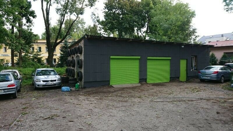 Szary budynek warsztatu z dwoma zielonymi bramami rolowanymi, opony zawieszone na ścianie, zaparkowane samochody przed budynkiem, teren częściowo utwardzony, częściowo ziemny.