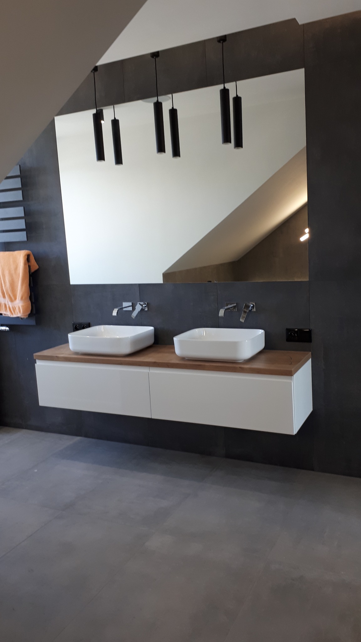 Nowoczesna łazienka na poddaszu z dwoma kwadratowymi umywalkami nablatowymi, drewnianym blatem, białymi szafkami pod umywalkami, czarną ścianą, dużym lustrem i designerskimi lampami wiszącymi.