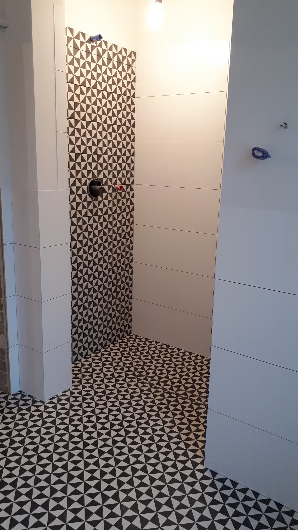 Wnętrze łazienki w trakcie remontu, widoczne białe płytki na ścianach, fragment ściany z wzorem czarno-białych trójkątów oraz podłoga wyłożona czarno-białymi płytkami w trójkąty, surowy otwór...