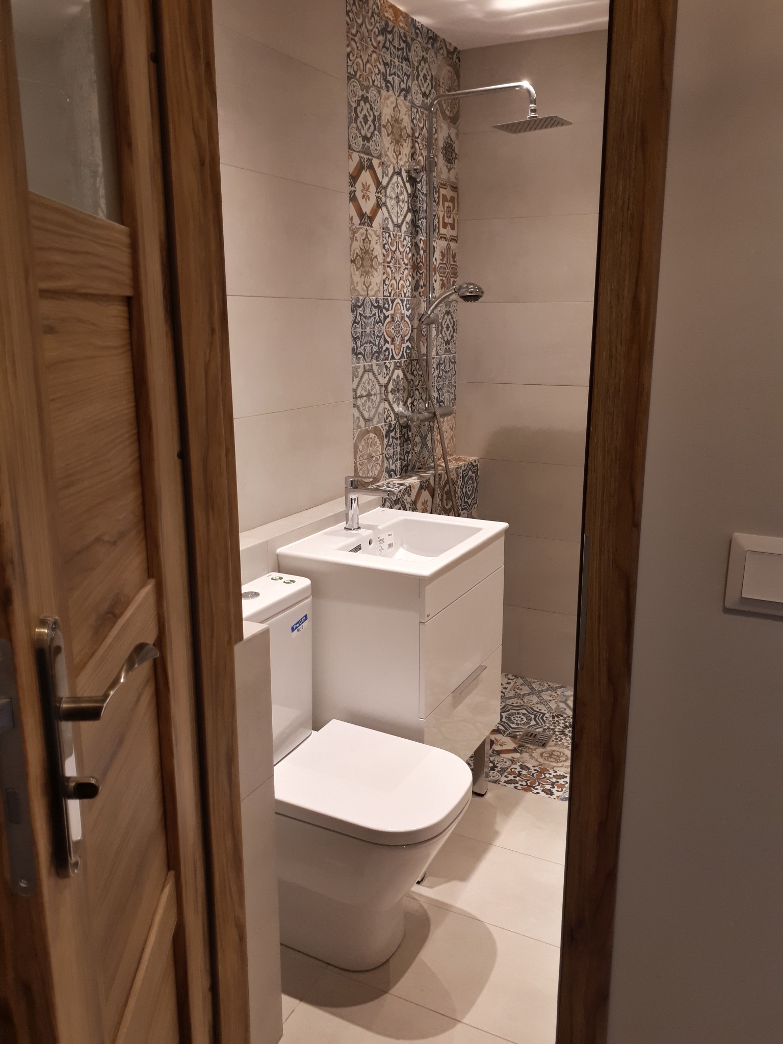 Nowoczesna łazienka z białą armaturą, wzorzystymi płytkami pod prysznicem i na podłodze, widziana zza drewnianych drzwi.