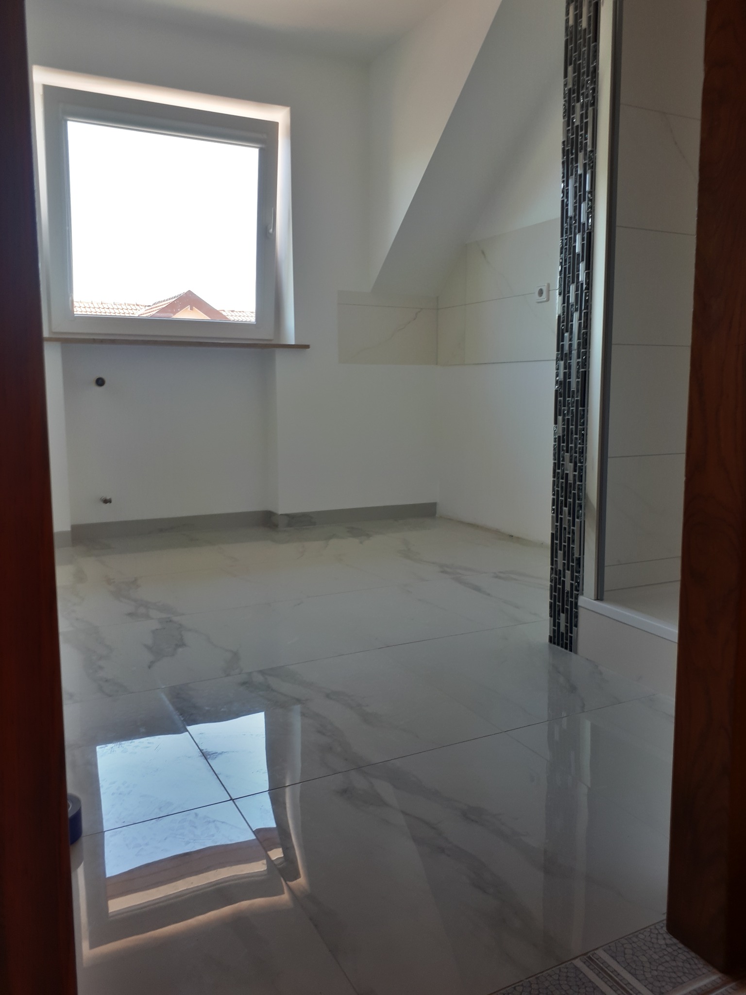 Nowa łazienka w trakcie wykańczania, widoczne błyszczące płytki podłogowe imitujące marmur, białe ściany, okno z widokiem na dach oraz fragment kabiny prysznicowej z dekoracyjną mozaiką.