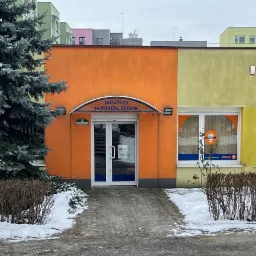 Fasada budynku biurowego pomalowana na pomarańczowo i żółto, z logo Osram w oknie i szyldem 'Biuro Handlowe' nad wejściem, otoczona zielenią i resztkami śniegu.