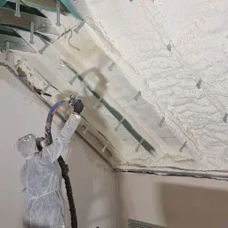 Pracownik w kombinezonie ochronnym natryskuje pianę izolacyjną na poddasze z widocznymi elementami konstrukcyjnymi dachu i specjalnymi uchwytami montażowymi.