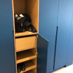 Niebieska zabudowa meblowa z otwartą szafką, widoczne półki z butami, szuflada i górna przestrzeń z torbą i ubraniami, zabezpieczona podłoga.