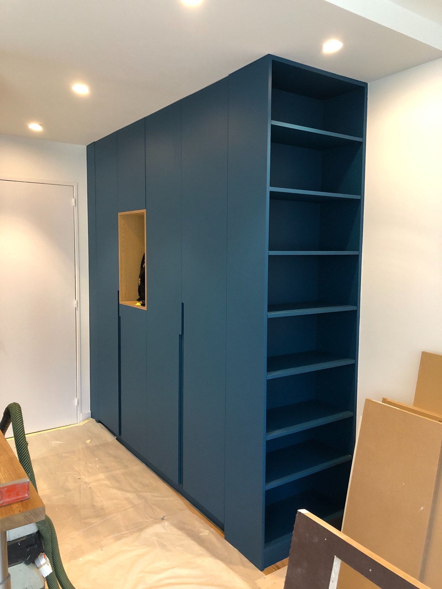 Niebieska zabudowa meblowa z szafkami i regałem z półkami, widoczne zabezpieczenia podłogi folią malarską, fragment odkurzacza przemysłowego i płyty meblowe.