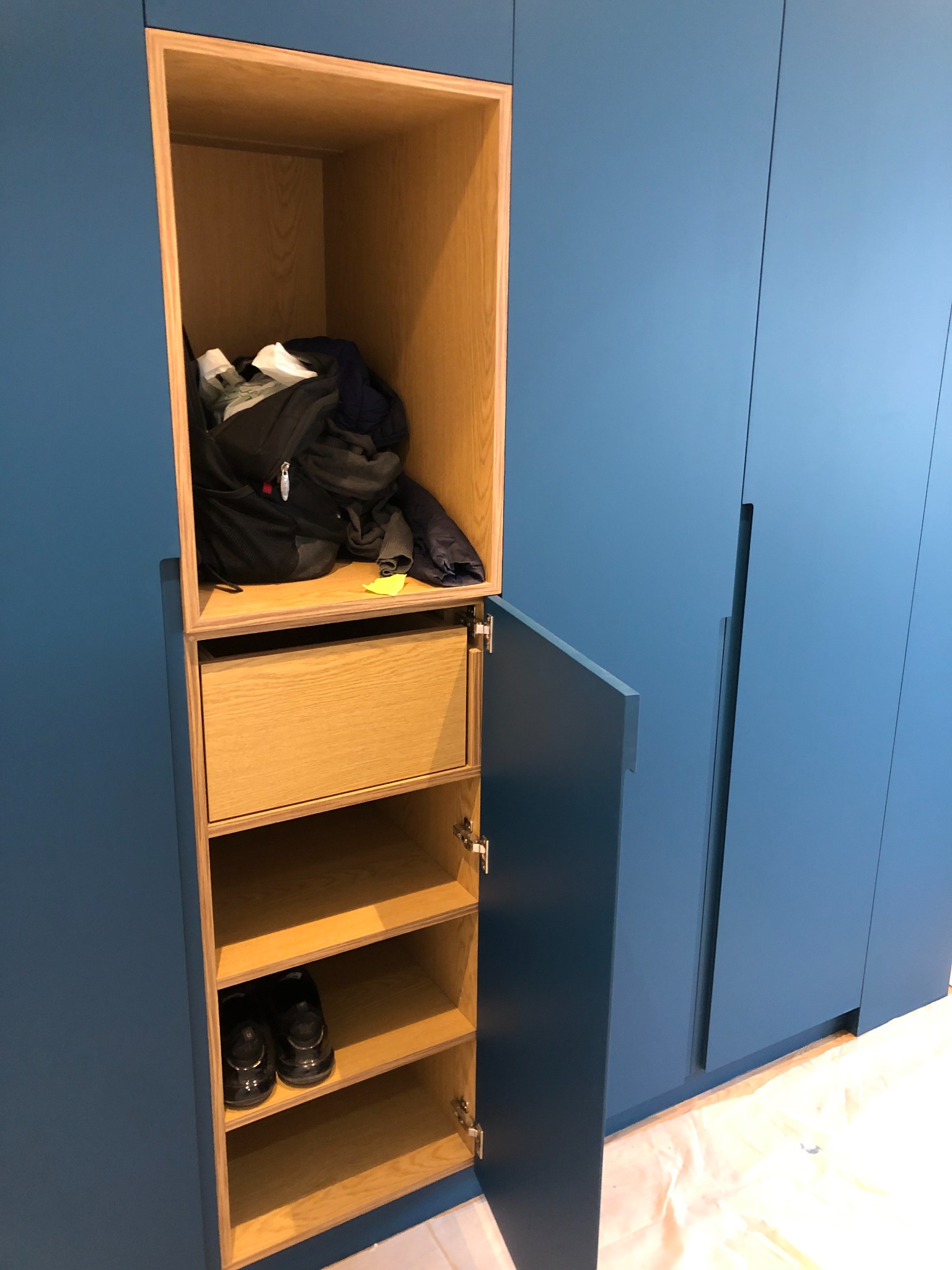 Niebieska zabudowa meblowa z otwartą szafką, widoczne półki z butami, szuflada i górna przestrzeń z torbą i ubraniami, zabezpieczona podłoga.