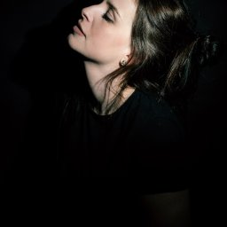 Magdalena Białek - Portret kobiety z profilu, oświetlona punktowo, z włosami upiętymi w kok i kolczykiem w kształcie kwiatka.