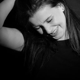 Magdalena Białek - Czarno-biały portret uśmiechniętej kobiety z długimi włosami, z ręką uniesioną do góry, widoczny kolczyk w kształcie kwiatka.