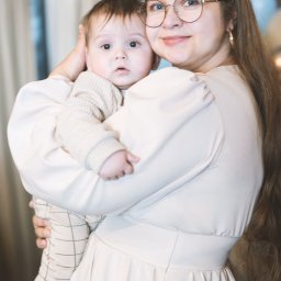 Magdalena Białek - Uśmiechnięta kobieta w okularach obejmuje małe dziecko ubrane w sweterek i spodnie w kratę na tle jasnych balonów.