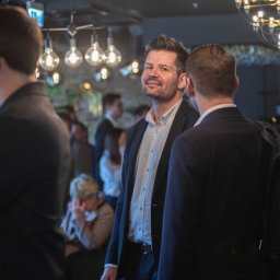 Magdalena Białek - Mężczyzna w garniturze uśmiecha się, patrząc w obiektyw podczas eventu firmowego, w tle widoczni inni uczestnicy i dekoracyjne oświetlenie.