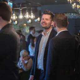 Mężczyzna w garniturze uśmiecha się, patrząc w obiektyw podczas eventu firmowego, w tle widoczni inni uczestnicy i dekoracyjne oświetlenie.