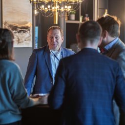 Magdalena Białek - Mężczyzna w niebieskiej marynarce rozmawia z grupą osób podczas wydarzenia w pomieszczeniu z nowoczesnym oświetleniem i ciemnym wystrojem.