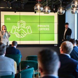 Magdalena Białek - Prezentacja z ekranem wyświetlającym zielony slajd z napisem 'PRIVATE OFFERS' i grafiką uścisku dłoni z tarczą, kobieta w marynarce w kratę przemawia do publiczności, mężczyzna w garniturze stoi...